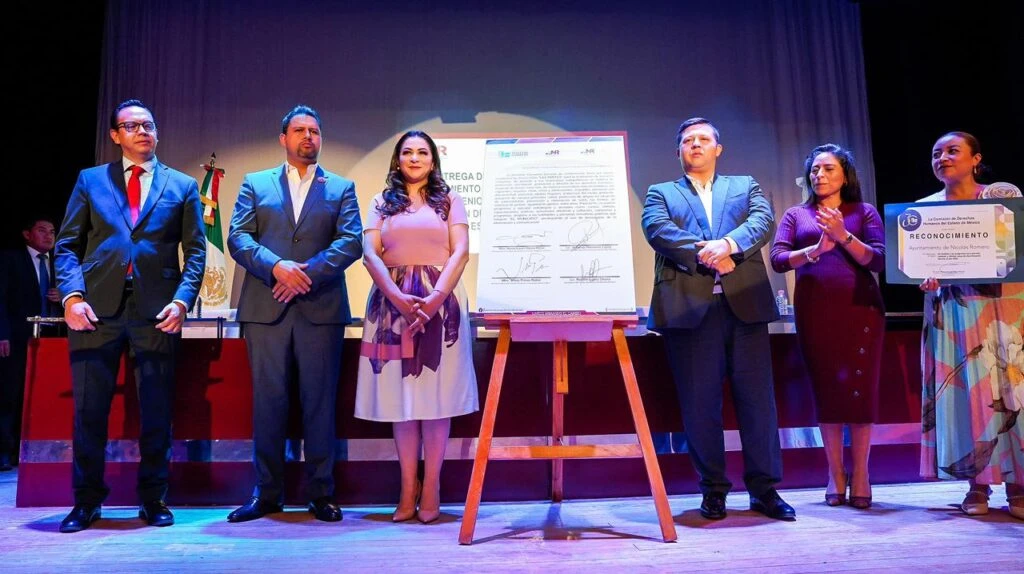 Grupo de seis personas alineadas sobre un escenario durante una ceremonia institucional, con un documento firmado en tamaño grande colocado en un soporte al centro; una bandera y mobiliario oficial se encuentran al fondo.