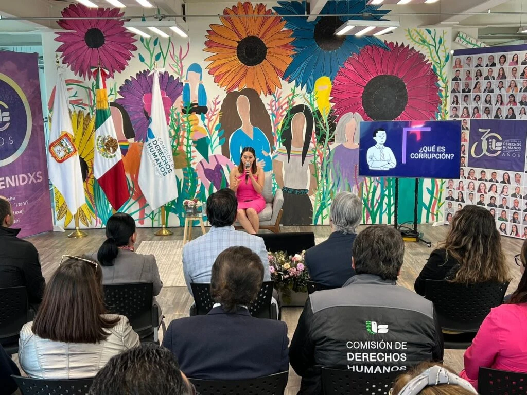 Sala de conferencias institucional con público sentado en sillas frente a una ponente ubicada al centro, quien habla desde un sillón individual; al fondo hay un mural colorido con flores grandes y figuras humanas, banderas oficiales y pantallas con información del evento.