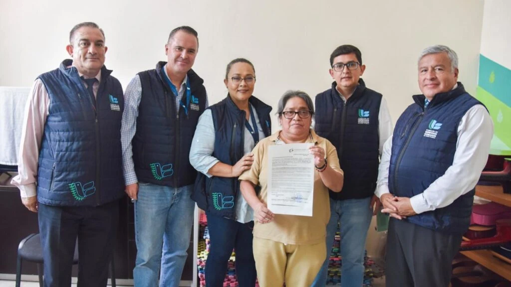 Grupo de seis personas con chalecos institucionales posan en interior junto a una persona que sostiene un documento oficial; el entorno incluye mobiliario y materiales en segundo plano dentro de una sala.