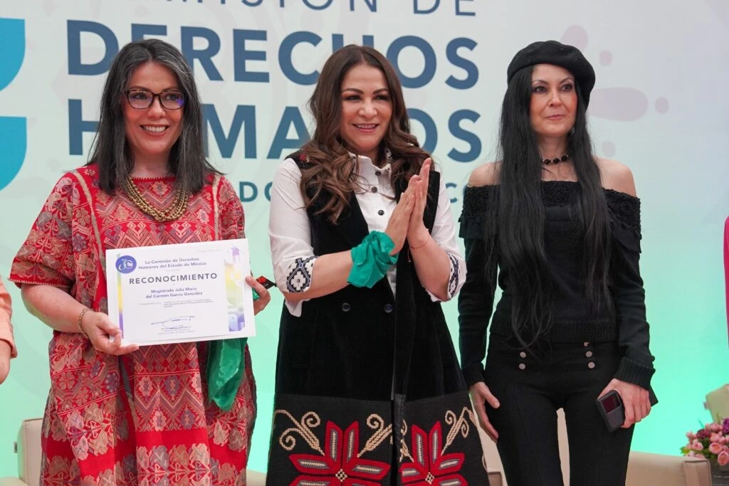 Tres personas de pie en un escenario institucional; una sostiene un reconocimiento enmarcado mientras las otras aplauden, con fondo de logotipo de la Comisión de Derechos Humanos y decoración formal.