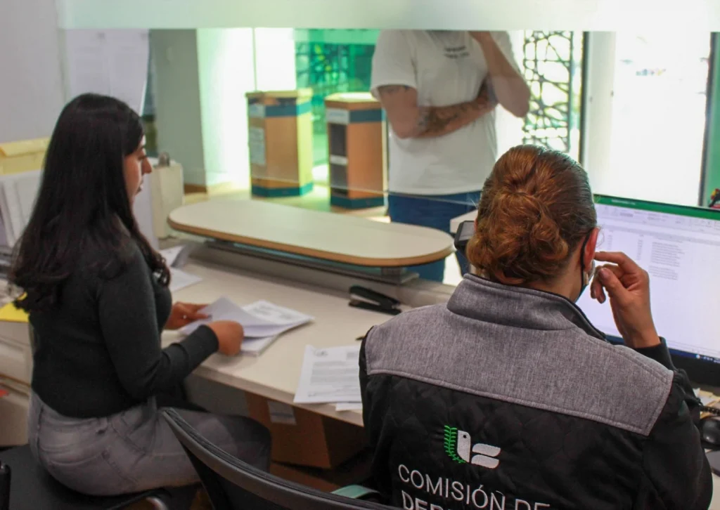 Área de atención en oficina donde dos personas se encuentran sentadas detrás de un mostrador revisando documentos y una computadora; frente a ellas, otra persona permanece de pie realizando un trámite. El espacio cuenta con ventanilla de servicio, mobiliario de oficina y luz natural proveniente de una ventana con protección.