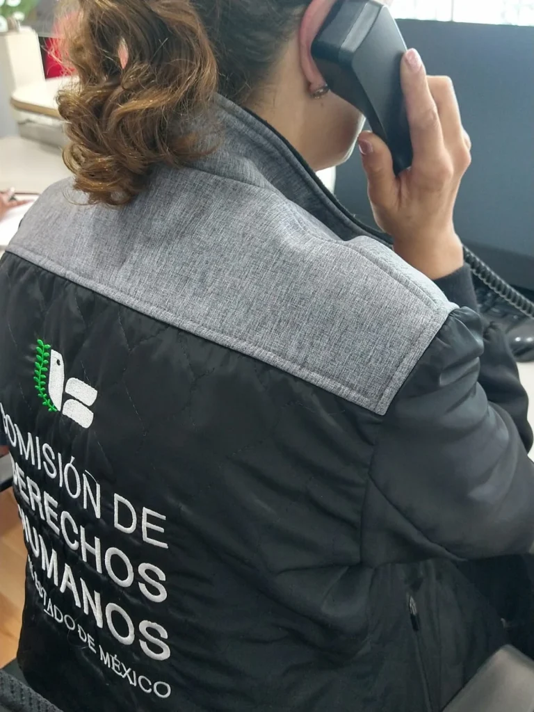 Primer plano de una persona en una estación de trabajo de oficina utilizando un teléfono fijo, vista desde atrás; viste un chaleco negro con el logotipo de la Comisión de Derechos Humanos del Estado de México, mientras frente a la persona se observa equipo de cómputo y mobiliario de oficina en un entorno administrativo.
