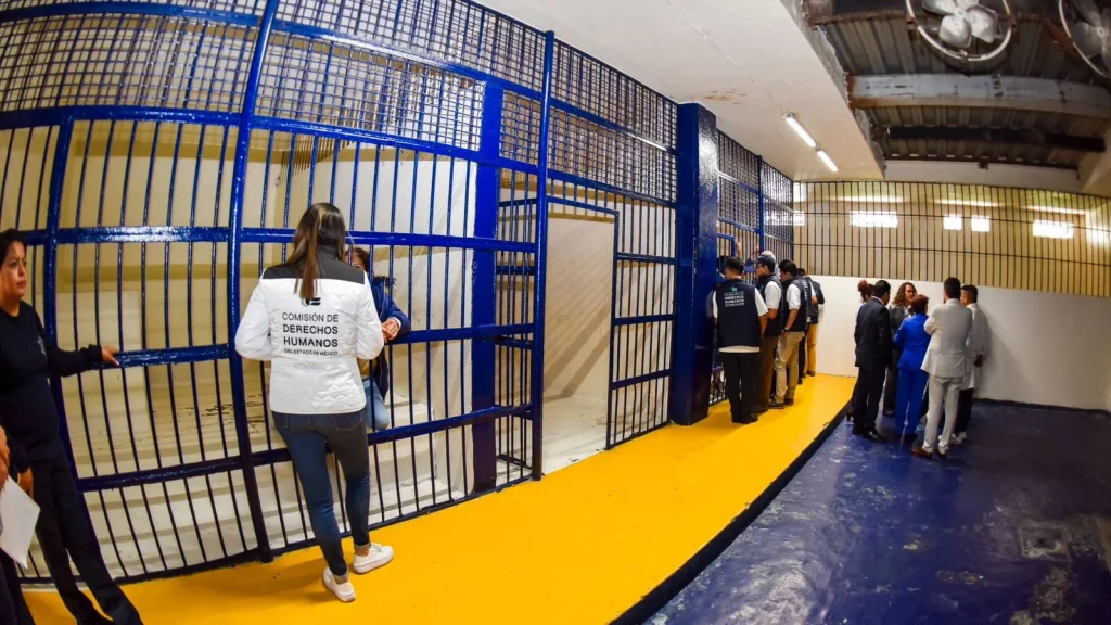 “Personal de la Comisión de Derechos Humanos del Estado de México realiza una inspección en una zona de celdas con barrotes azules dentro de un centro de detención, mientras un grupo de personas observa desde un pasillo con piso amarillo y azul.”