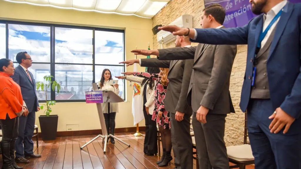Ceremonia oficial en una sala institucional: varias personas de pie con el brazo extendido realizan un juramento frente a un podio donde una persona lee un documento. El espacio cuenta con ventanas grandes, piso de madera, una bandera y un letrero morado de fondo.