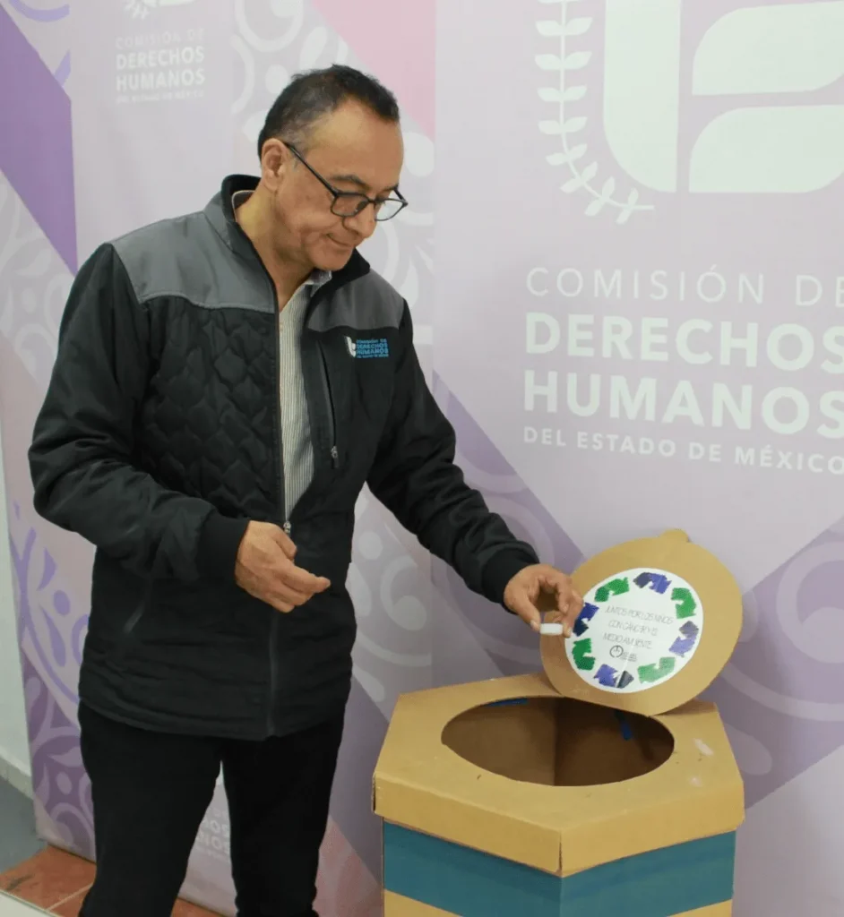 Persona con gafas y chamarra oscura deposita un objeto pequeño en un contenedor circular de reciclaje hecho de cartón, frente a un fondo institucional con el texto “Comisión de Derechos Humanos del Estado de México” y gráficos decorativos.