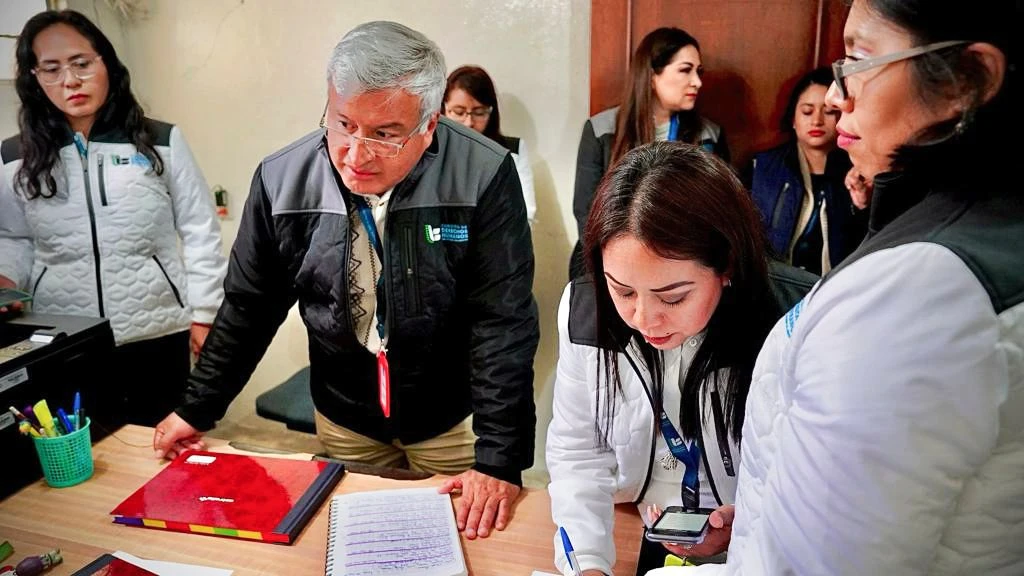 Grupo de varias personas con chalecos institucionales reunidas alrededor de un escritorio en una oficina; una persona escribe en un cuaderno mientras otra observa de cerca y otras permanecen alrededor, junto a objetos como carpetas, un portaplumas y equipo de oficina.