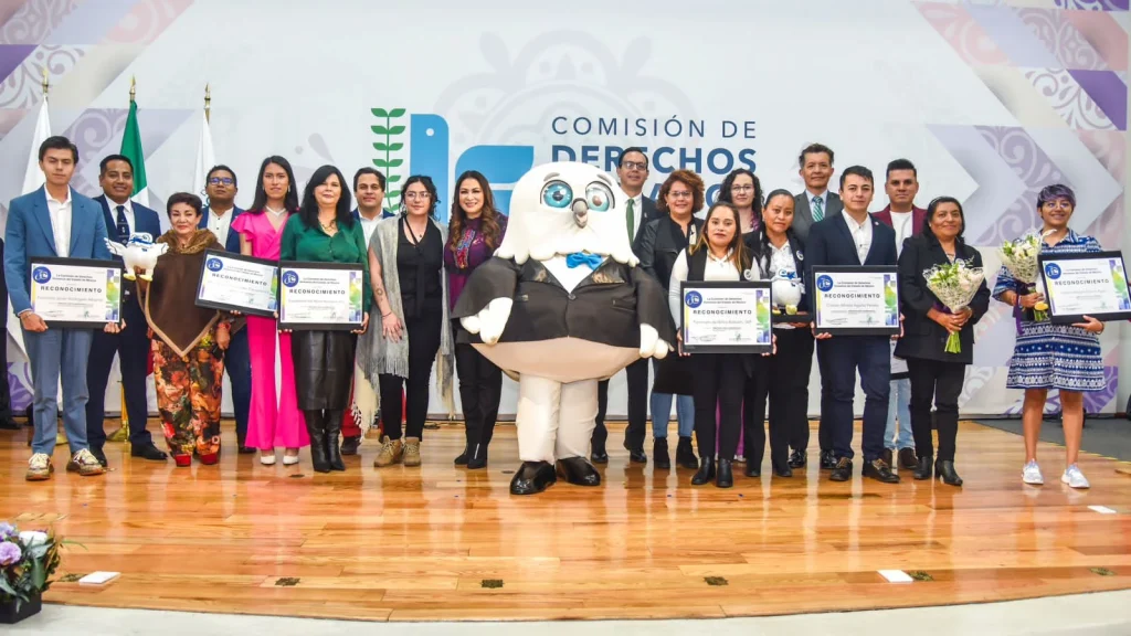 Grupo numeroso de personas posando sobre un escenario de piso de madera durante una ceremonia institucional, frente a un fondo con el logotipo y texto “Comisión de Derechos Humanos”. Varias personas sostienen diplomas de reconocimiento enmarcados, en compañía de Jeasu, símbolo de la CODHEM con traje formal se encuentra al centro. También se observan banderas y arreglos florales en los extremos del escenario.