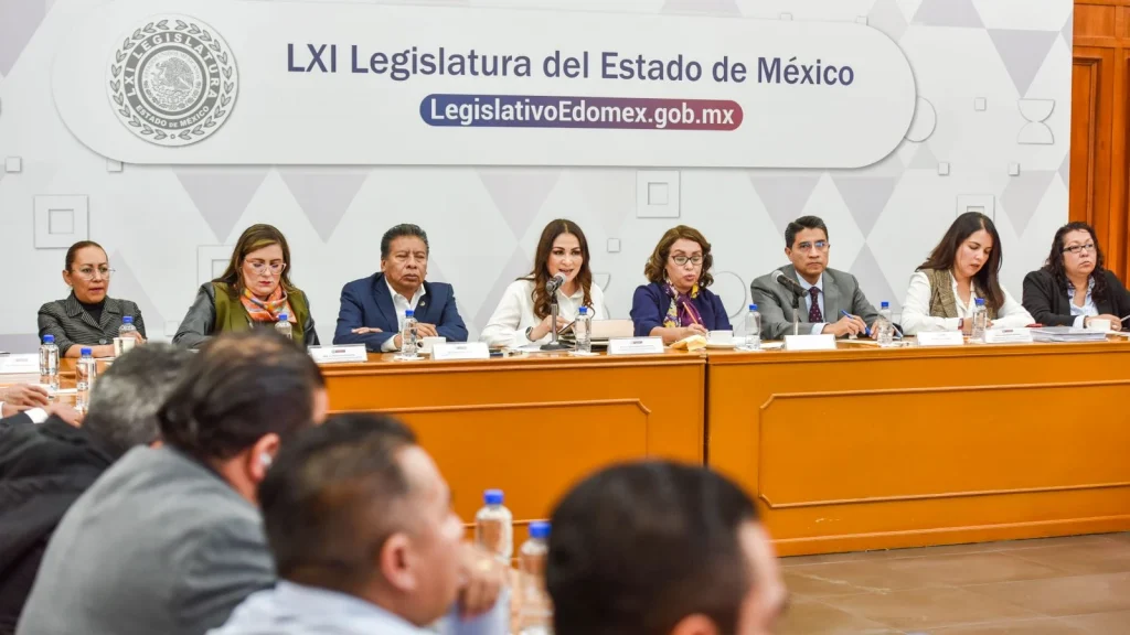 Mesa de trabajo en una sala institucional donde varias personas se encuentran sentadas detrás de un escritorio largo con micrófonos y botellas de agua, durante una sesión o reunión formal. Al fondo se observa el emblema y texto “LXI Legislatura del Estado de México” junto con la dirección “LegislativoEdomex.gob.mx”. En primer plano se distinguen asistentes sentados frente a la mesa principal.