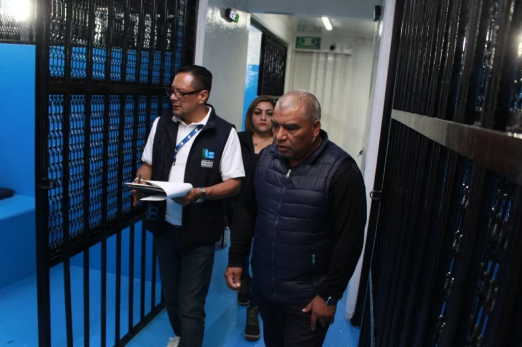 Pasillo interior con celdas metálicas a ambos lados en un espacio cerrado similar a un área de reclusión. Varias personas caminan por el pasillo; una de ellas porta un chaleco institucional y sostiene una libreta con documentos, mientras otras acompañan. El área está iluminada con luz artificial y presenta paredes blancas con partes en color azul, rejas negras y señalización de salida al fondo.