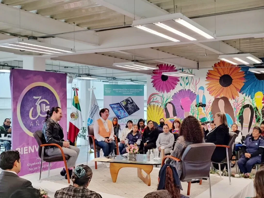Espacio interior utilizado para una conversación o panel, con varias personas sentadas en semicírculo alrededor de una mesa de centro con flores. En el área se observan sillas individuales y público asistente. El fondo incluye un mural colorido con flores