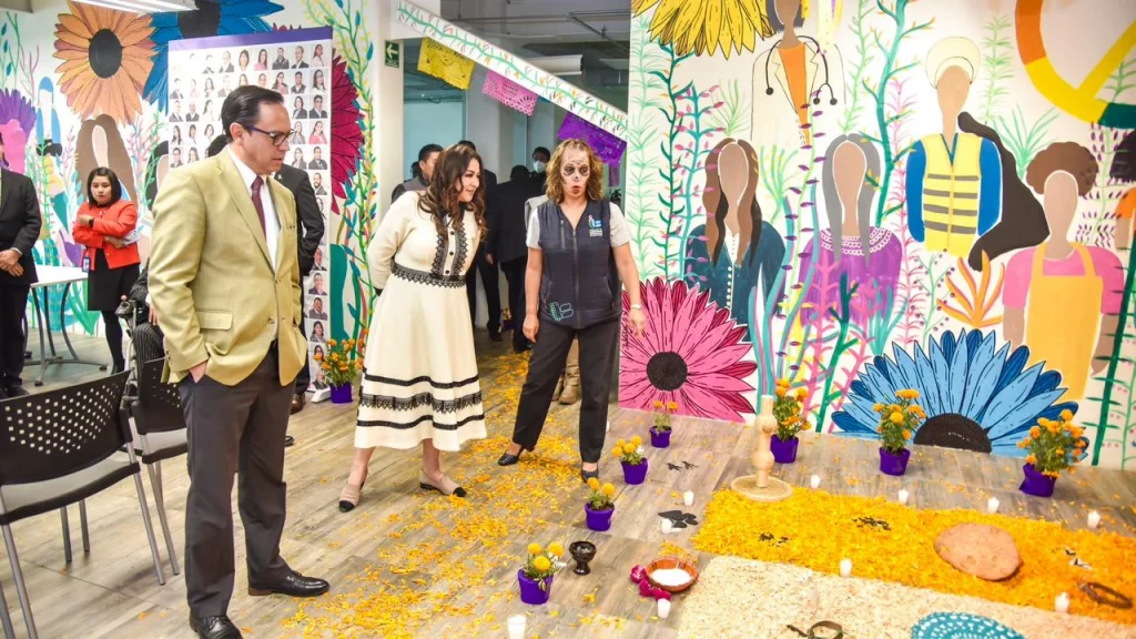 Espacio interior decorado con un mural colorido de flores grandes y figuras humanas estilizadas, donde varias personas observan una ofrenda tradicional colocada en el suelo. La ofrenda incluye pétalos de flor de cempasúchil, veladoras, pequeños recipientes y objetos simbólicos. En el área también se ven macetas con flores, sillas alrededor y un ambiente preparado para una actividad cultural o conmemorativa dentro de un recinto institucional.