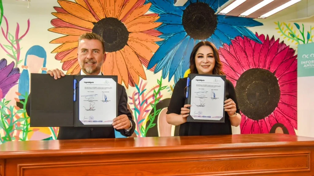 Dos personas se encuentran detrás de una mesa de madera en un espacio interior, sosteniendo carpetas abiertas con documentos oficiales firmados. Al fondo se observa un mural colorido con flores de gran tamaño en tonos naranja, azul y rosa, acompañado de elementos decorativos vegetales. La escena incluye iluminación de techo con paneles rectangulares y un entorno institucional o de presentación pública.
