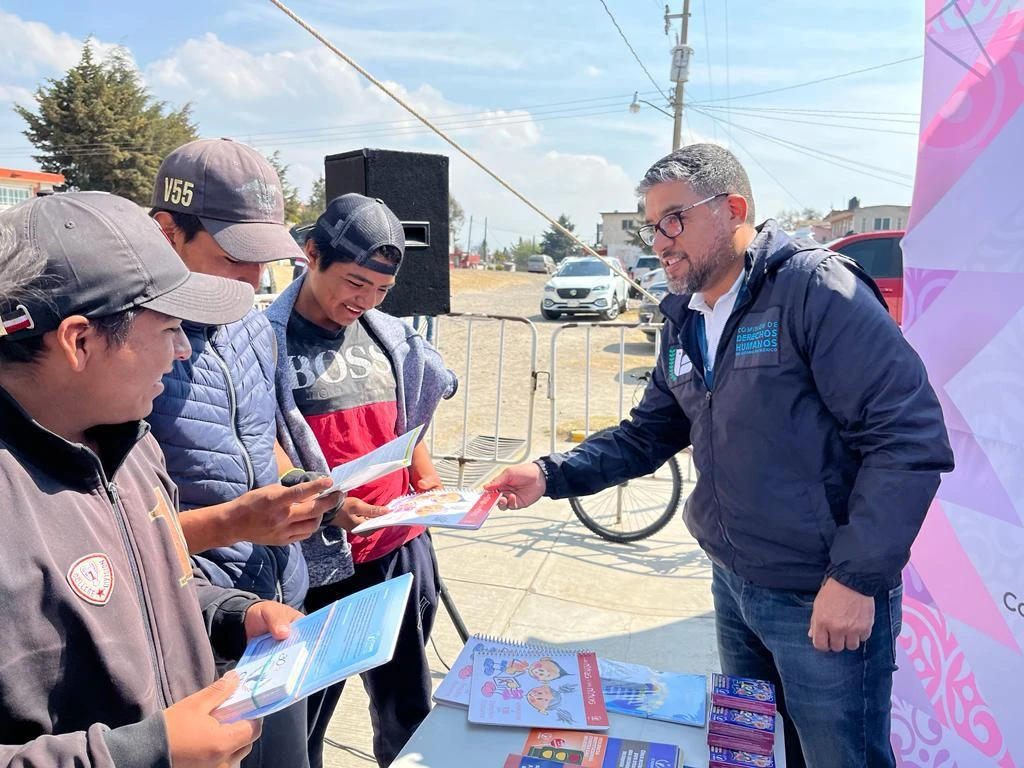 Actividad informativa realizada en una calle al aire libre, donde una persona con chamarra institucional entrega materiales impresos a tres personas jóvenes que sostienen folletos y los revisan. En primer plano se observa una mesa con más materiales de difusión, mientras que en el fondo hay vehículos, una bocina, una bicicleta y viviendas, en un entorno urbano con cielo despejado.