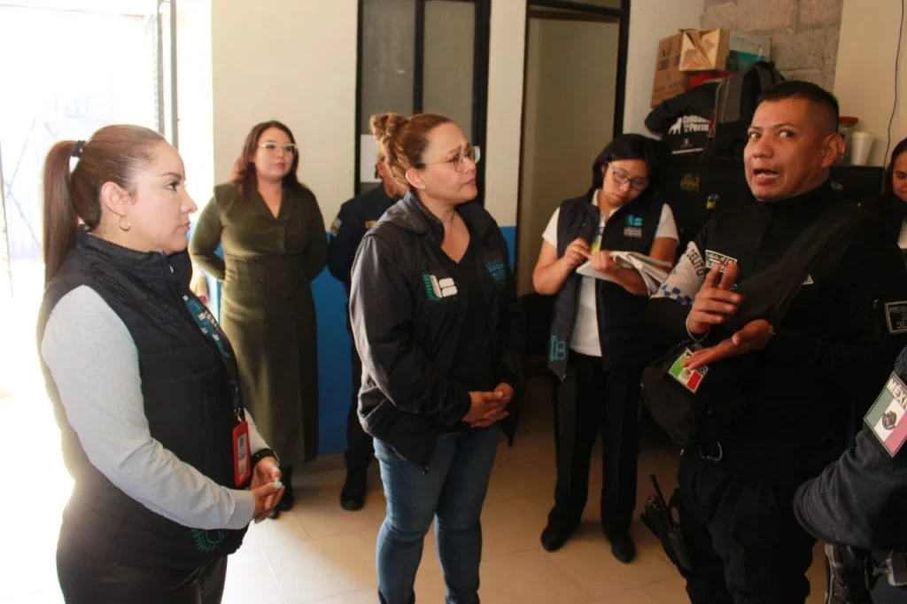 Espacio interior donde un grupo de varias personas se encuentra de pie participando en una actividad de supervisión o diálogo institucional. Al frente, una persona con uniforme oscuro y distintivos oficiales habla y gesticula mientras explica algo al grupo. Varias personas alrededor, algunas con chalecos institucionales y credenciales, escuchan y toman notas en una libreta. El lugar tiene paredes con colores claros y azul, puertas de acceso y mobiliario básico, lo que sugiere un entorno administrativo o de servicio.