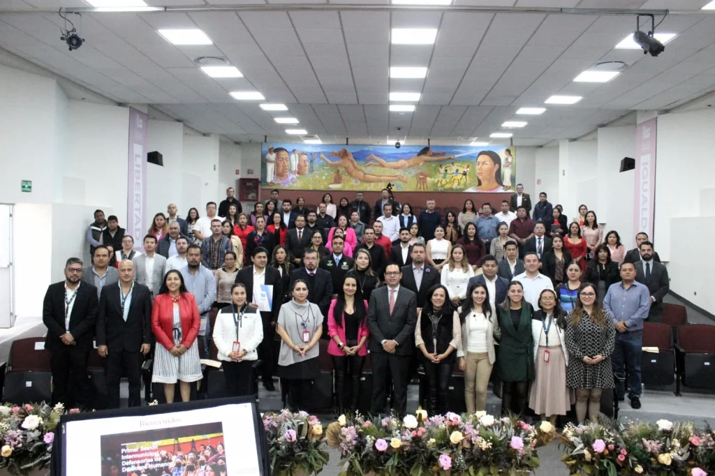 Fotografía grupal en un auditorio donde decenas de personas se encuentran de pie organizadas en filas frente a asientos escalonados. En la parte frontal hay un arreglo floral sobre una mesa y una pantalla con la palabra “Bienvenidos” y referencia a una “Primer Sesión Intermunicipal de Defensorías de Derechos Humanos”.