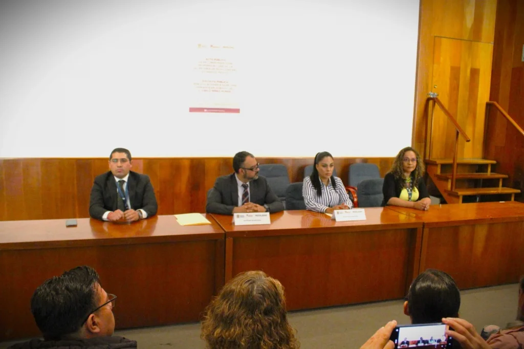 Sala de conferencias con una mesa larga de madera al frente, donde cuatro personas están sentadas como panel durante una actividad formal. Detrás de la mesa hay una pared blanca con proyección de texto. El espacio cuenta con iluminación de plafones en el techo y escaleras laterales de acceso, lo que sugiere un entorno institucional o académico para una presentación pública.