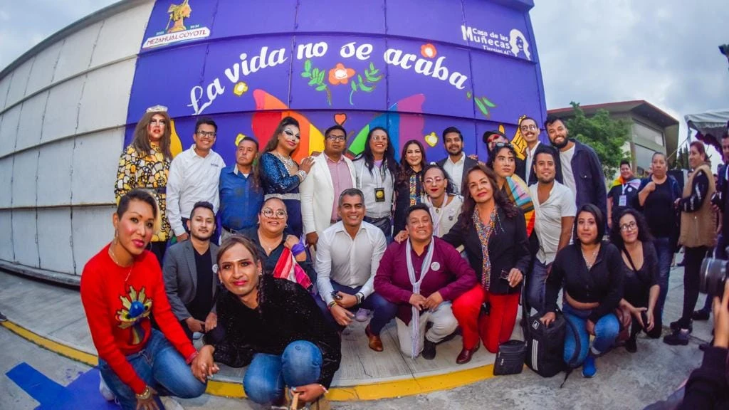 Grupo numeroso de personas posando al aire libre frente a un muro morado con un mural colorido y la frase “La vida no se acaba”. Varias figuras están de pie y otras en cuclillas en la parte frontal, organizadas en filas para la fotografía. El fondo incluye ilustraciones de flores, un arco multicolor y logotipos institucionales, mientras que alrededor se observan edificios, cielo nublado y elementos de una calle o explanada.