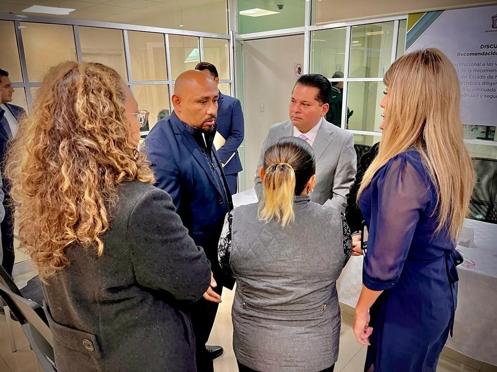 Oficina con paredes de cristal y marcos blancos donde varias personas forman un círculo mientras conversan. En el centro, dos personas con vestimenta formal dialogan frente a frente, mientras otra persona con uniforme oscuro participa activamente.