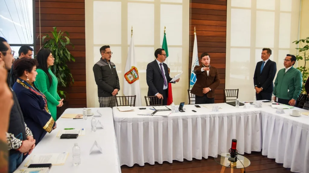Sala de reuniones institucional con mesas cubiertas con manteles blancos dispuestas en forma de U, donde varias personas están de pie alrededor durante una sesión formal. En el centro, una persona sostiene documentos y otra realiza un gesto con el brazo extendido, como parte de una intervención o toma de protesta