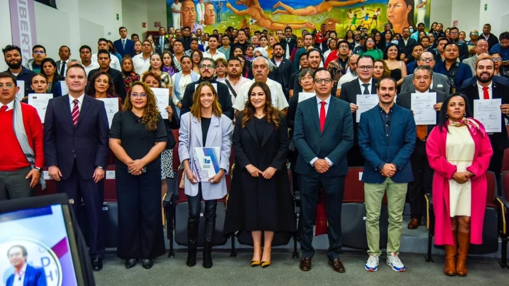 Fotografía grupal en un auditorio con asientos escalonados, donde un gran número de personas se encuentra de pie organizada en varias filas. En la primera fila, varias personas sostienen documentos o diplomas y portan vestimenta formal