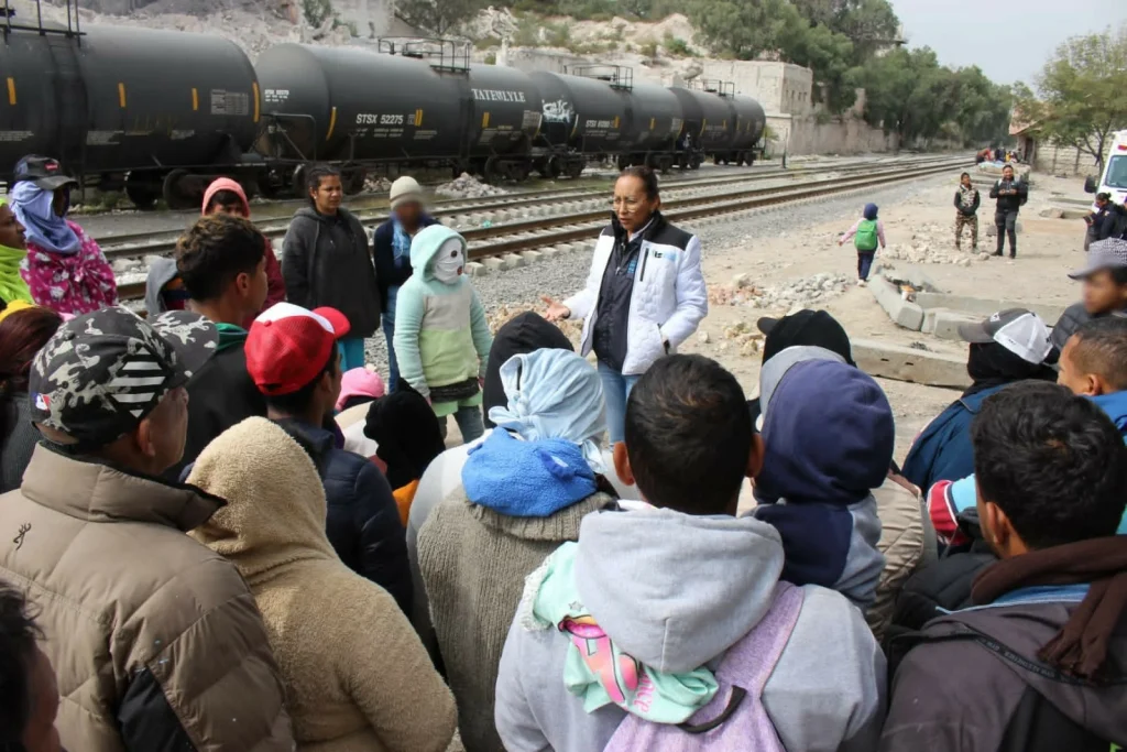 Grupo de personas reunidas al aire libre junto a vías de tren, donde una persona con chaleco institucional y chamarra clara explica información a un conjunto de personas que la rodean. En primer plano, varias personas observan y escuchan, algunas con mochilas o ropa abrigadora. Al fondo se ven vagones de carga