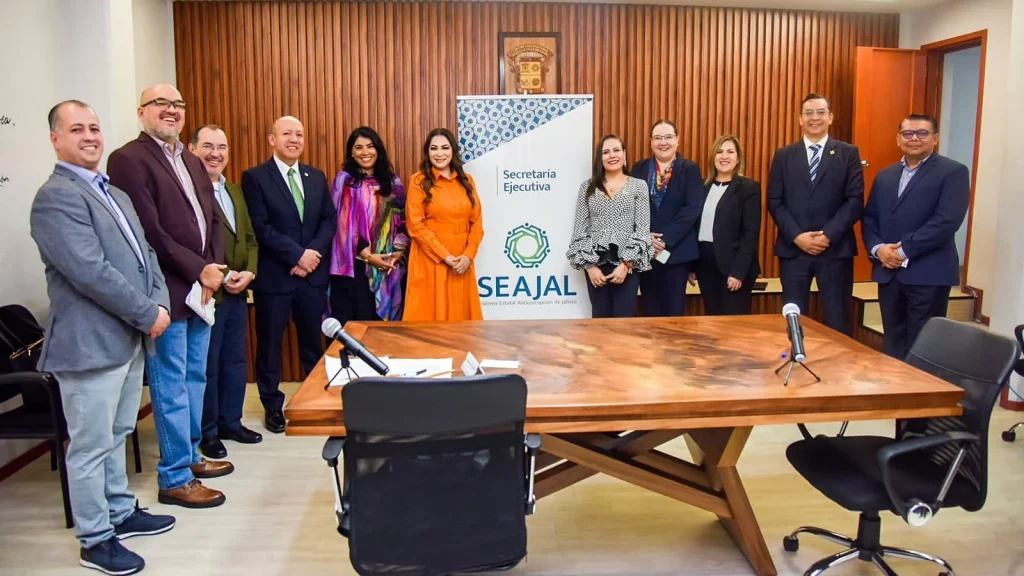Grupo de personas posando en una sala de reuniones con paneles de madera en la pared, detrás de una mesa rectangular de madera con micrófonos. Al fondo hay un estandarte con el texto “Secretaría Ejecutiva” y el logotipo de “SEAJAL (Sistema Estatal Anticorrupción de Jalisco)”. Las personas están alineadas frente a la mesa en un ambiente institucional bien iluminado.