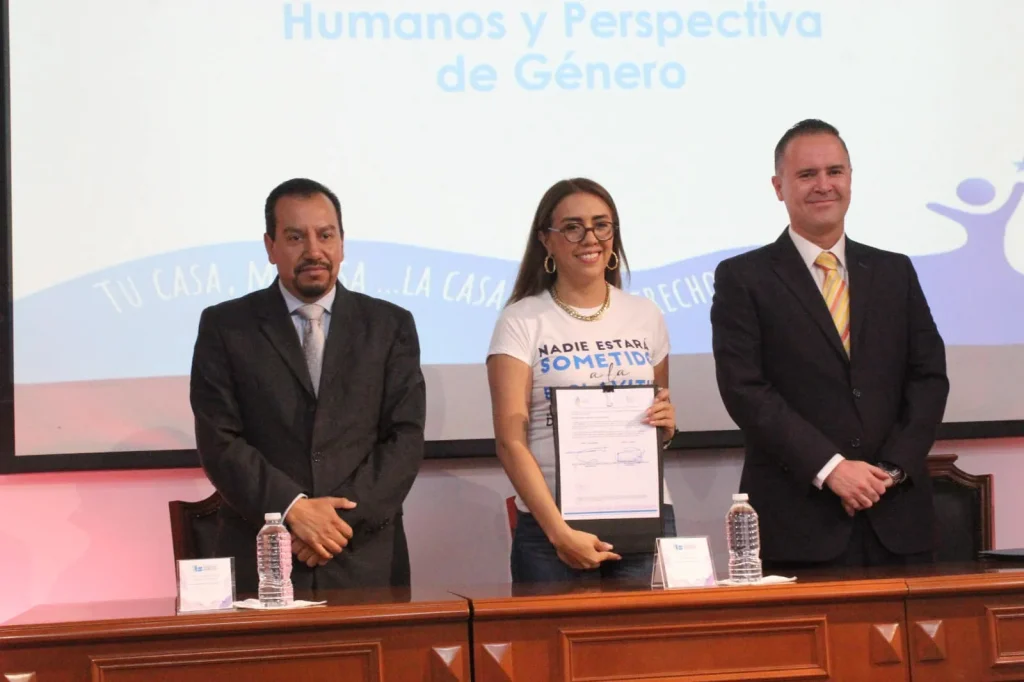 Tres personas posan detrás de una mesa larga de madera en un auditorio o sala institucional. En el centro, una persona sostiene un documento enmarcado. Sobre la mesa hay botellas de agua y tarjetas de identificación. En el fondo se proyecta una pantalla con el texto “Humanos y Perspectiva de Género” y otros elementos gráficos en tonos claros.