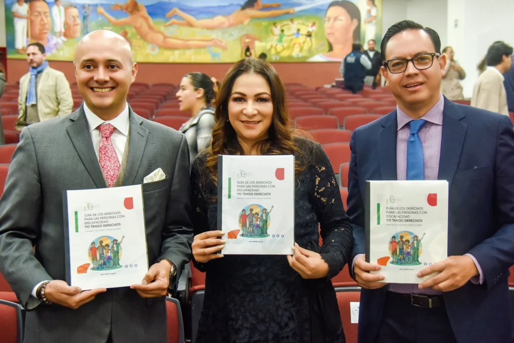 Tres personas posan en un auditorio con filas de asientos rojos al fondo. Cada una sostiene una publicación titulada “Guía de los derechos para las personas con discapacidad: yo tengo derechos”, que incluye una ilustración de figuras humanas. Detrás se observa un mural colorido con figuras humanas y escenas simbólicas, así como otras personas de pie en el espacio.