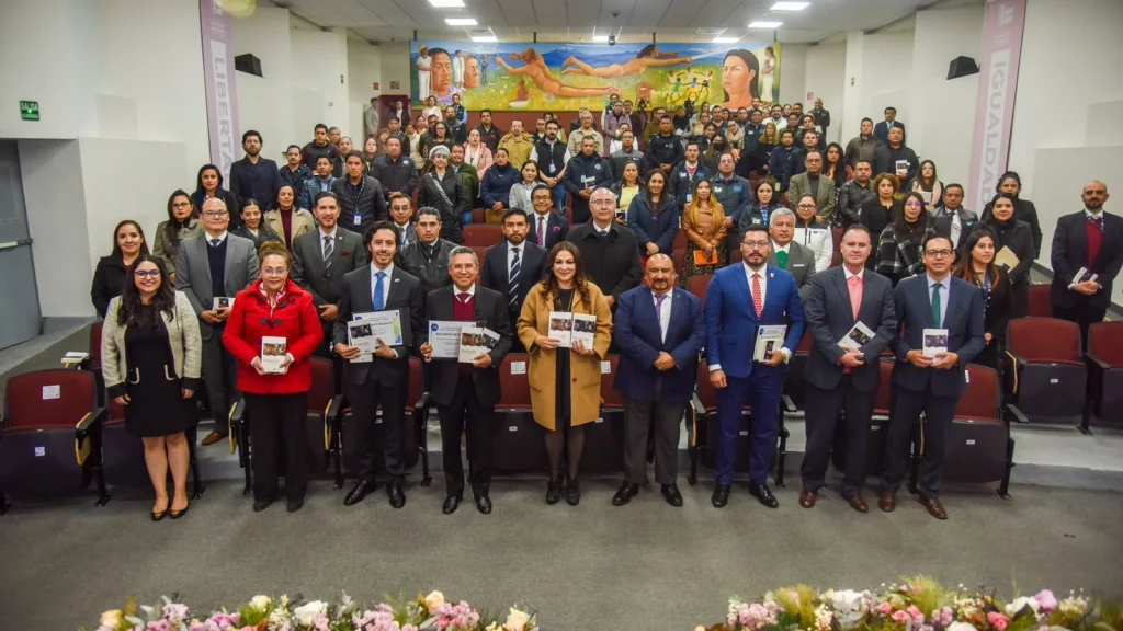 Grupo numeroso de personas reunidas en un auditorio con asientos rojos dispuestos en filas. En primer plano, varias personas de pie sostienen publicaciones impresas, mientras detrás se observa una gran cantidad de asistentes también de pie en diferentes niveles del recinto