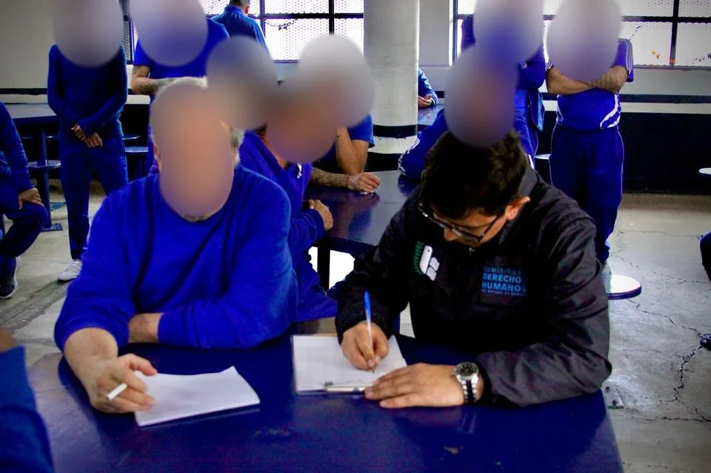 Varias personas vestidas con uniformes azules se encuentran en una sala interior con mesas metálicas y ventanas al fondo