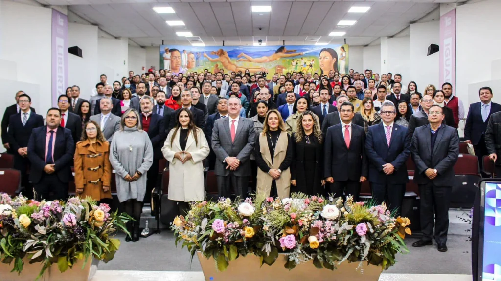 Grupo numeroso de personas en vestimenta formal posando en un auditorio interior, organizadas en varias filas frente a arreglos florales; al fondo se observa un mural colorido y en las paredes aparecen palabras como “IGUALDAD” y “LIBERTAD”.