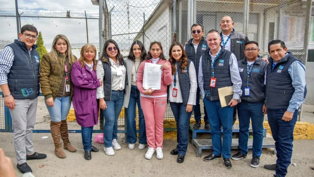 Grupo de aproximadamente once personas posando al aire libre frente a una cerca de malla metálica y una estructura institucional; varias personas llevan chalecos con logotipo y credenciales visibles, mientras una persona en el centro sostiene un documento impreso hacia la cámara.