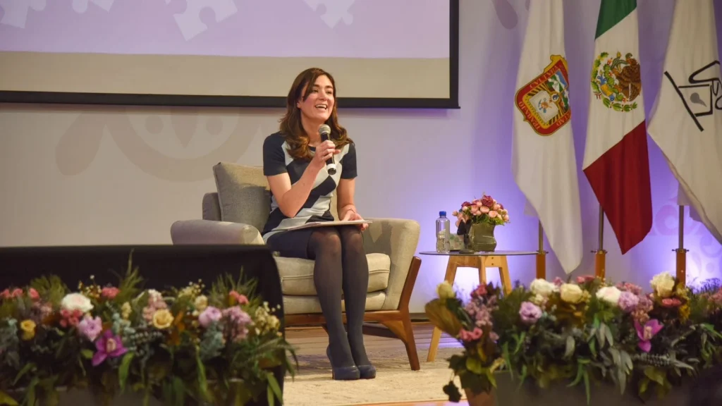 Persona sentada en un sillón sobre un escenario durante un evento público, sosteniendo un micrófono y un cuaderno. A un lado hay una mesa pequeña con un arreglo floral y una botella de agua.