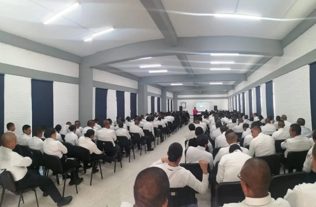 Sala de capacitación con personal policial sentado en sillas negras en filas ordenadas. Una persona con suéter rosa y credencial está de pie en el pasillo central, aparentemente impartiendo una charla. Al fondo se observa una pantalla de proyección, altavoces y un emblema en la pared, mientras la iluminación es uniforme y clara.
