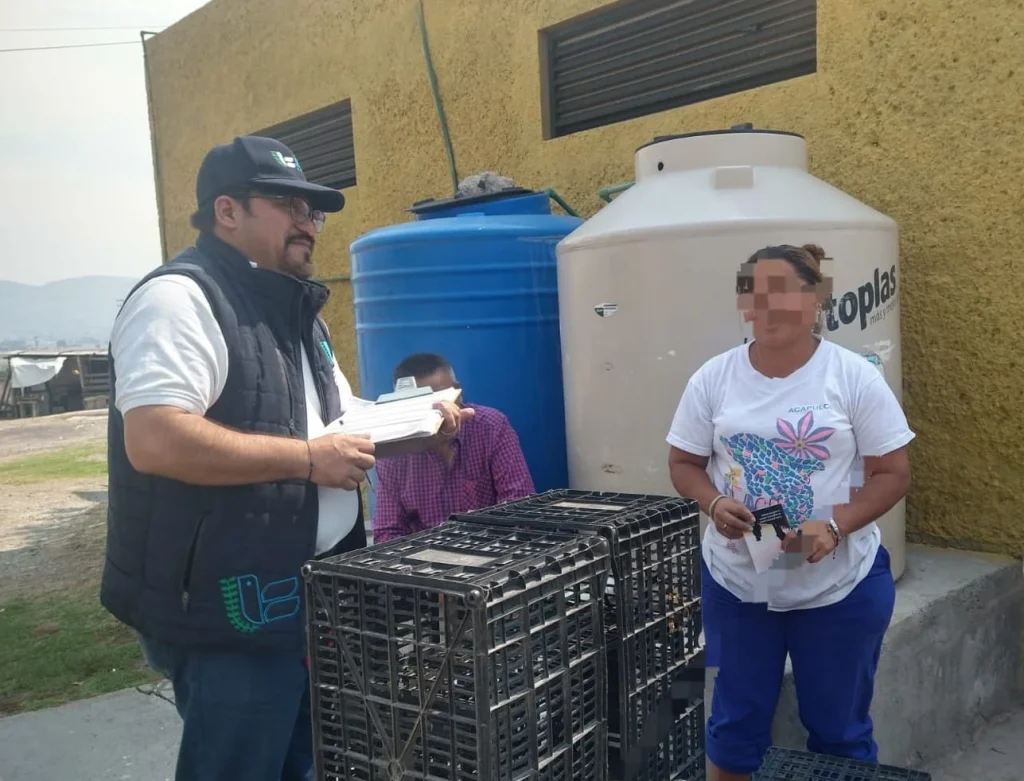 Dos personas al aire libre junto a un edificio con pared amarilla y contenedores de agua, una persona sostiene documentos frente a varias cajas plásticas negras apiladas mientras otra persona, con el rostro pixelado, sostiene un objeto pequeño; en el fondo se observan depósitos, una estructura rural y paisaje con montañas.