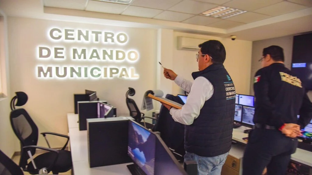 Interior de una sala de monitoreo con el letrero iluminado “CENTRO DE MANDO MUNICIPAL” en la pared; varias estaciones con computadoras y sillas están dispuestas en filas. En primer plano, una persona sostiene un portapapeles y señala hacia el frente, mientras otra persona camina dentro del área de trabajo.