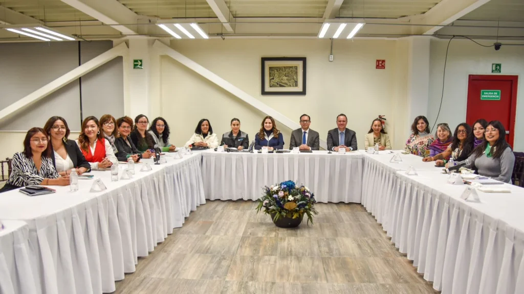 Grupo de personas sentadas alrededor de mesas dispuestas en forma de U durante una reunión formal en una sala interior, con un arreglo floral al centro y señalización de salida de emergencia en el fondo.