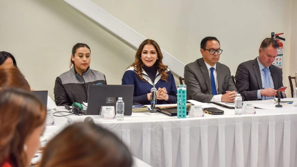 Grupo de personas participando en una sesión de trabajo sentadas detrás de una mesa con computadoras, micrófonos y botellas de agua, en un entorno interior tipo sala de juntas.