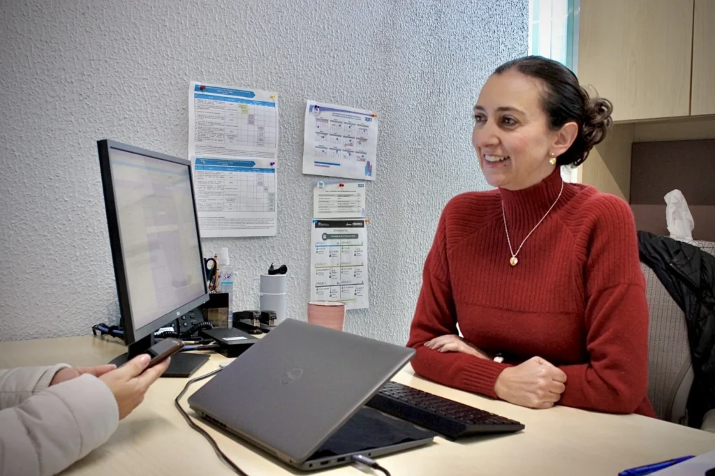 "Persona sentada frente a un escritorio sonriente mientras interactúa con otra persona fuera de cámara, con una computadora portátil y un monitor en un entorno de oficina."