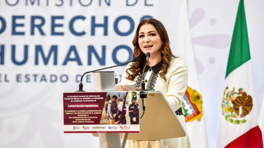 Persona hablando desde un podio con micrófono en un evento de la Comisión de Derechos Humanos del Estado de México. En el fondo se observa el logotipo institucional y a un lado la bandera de México. El podio tiene un cartel que menciona una capacitación presencial sobre administración pública e implementación de políticas municipales.