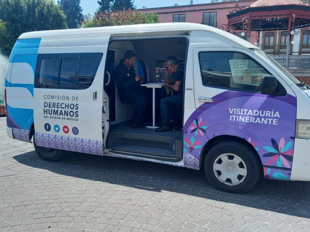 Unidad móvil de la Comisión de Derechos Humanos del Estado de México estacionada en una plaza. La puerta lateral del vehículo está abierta y en el interior se observa a dos personas sentadas frente a una pequeña mesa realizando una atención o trámite.