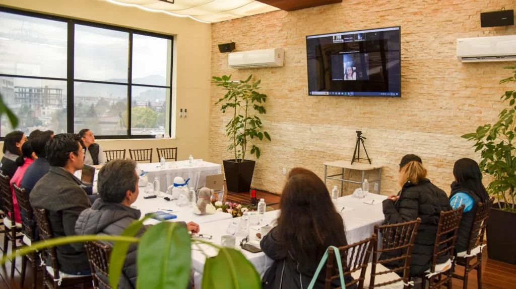 “Grupo de personas sentadas alrededor de mesas en una sala de reuniones con iluminación cálida, observando una videollamada en una pantalla montada en la pared; hay plantas, botellas de agua y equipo de trabajo sobre las mesas.”