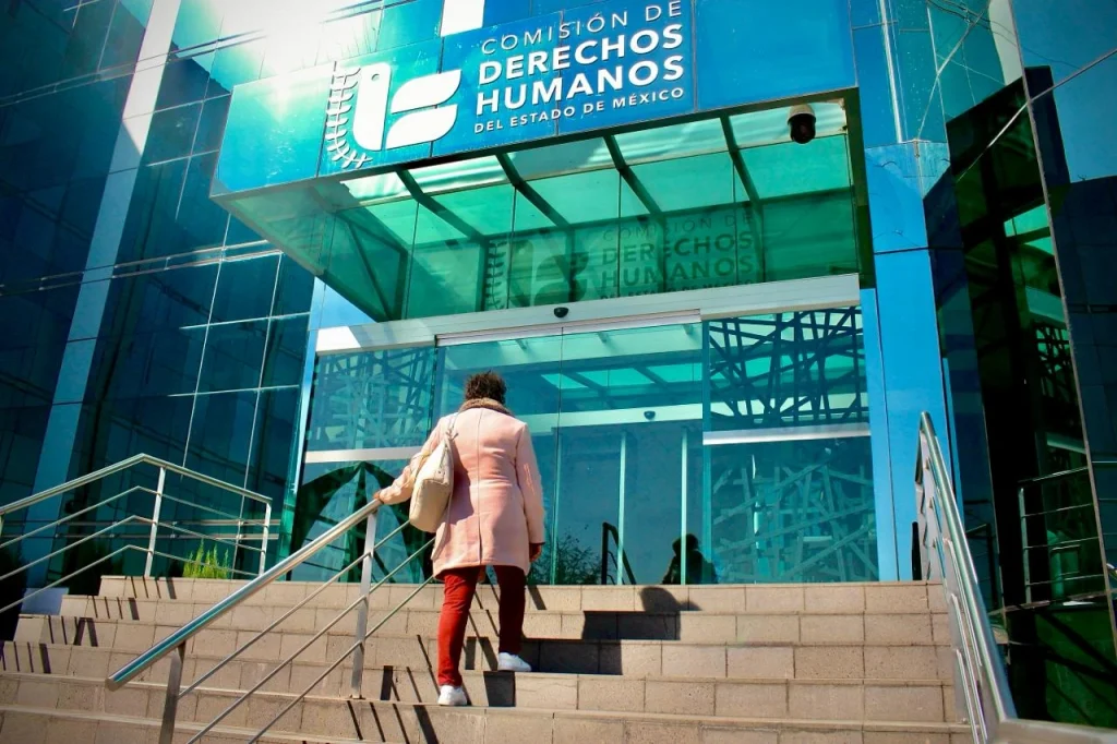 “Entrada de un edificio moderno con fachada de vidrio color azul que muestra el letrero ‘Comisión de Derechos Humanos del Estado de México’; una persona asciende por las escaleras hacia la puerta principal con barandales metálicos y un techo de cristal.”
