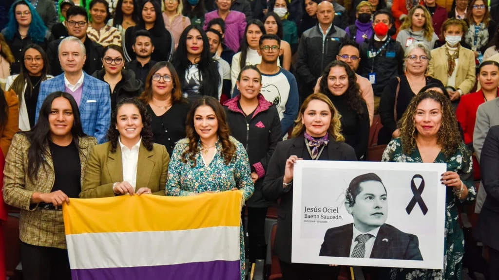 “Grupo numeroso de personas posando en un auditorio; en la primera fila varias sostienen una bandera de franjas amarilla, blanca y morada, y un cartel con el retrato y nombre ‘Jesús Ociel Baena Saucedo’ junto a un listón negro, mientras más asistentes aparecen al fondo en filas escalonadas.”