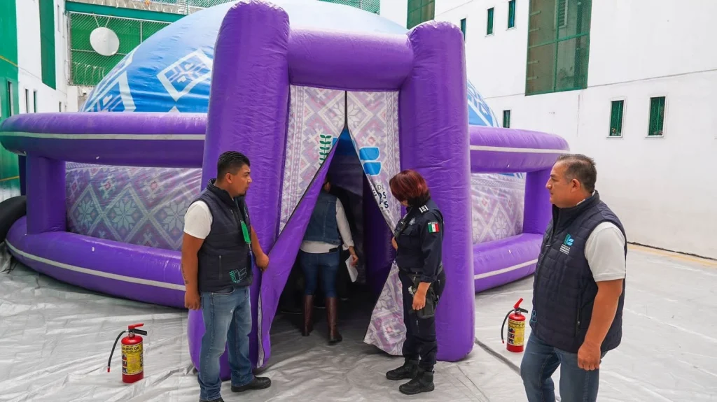 “Estructura inflable de color morado con forma de domo instalada en un patio, con una entrada abierta; varias personas se encuentran alrededor mientras una persona entra, y se observan dos extintores colocados a los lados de la entrada en el suelo.”