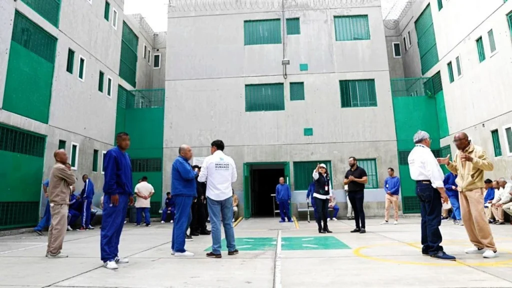 “Patio interior de un complejo con muros altos de concreto y ventanas con rejas verdes; varias personas vestidas con uniformes y ropa casual se encuentran distribuidas en el espacio, mientras otras personas conversan y observan frente a una entrada central del edificio.”