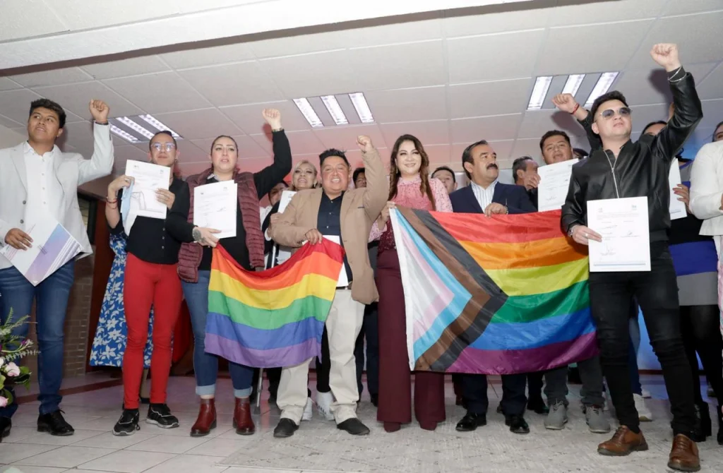 “Grupo de personas reunidas en un espacio interior mostrando documentos mientras levantan el brazo; al frente sostienen banderas multicolores, incluida una bandera arcoíris y otra con franjas adicionales, en un ambiente de evento o acto público.”