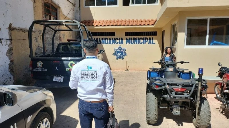 “Patio exterior de una comandancia municipal con paredes color beige y verde, donde varias personas se encuentran de pie; al frente hay vehículos oficiales, incluyendo una camioneta y un vehículo todo terreno, mientras una persona camina hacia el edificio con el logotipo de derechos humanos en la espalda.”
