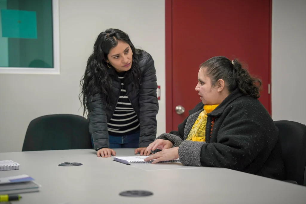 “Dos personas en una sala de oficina se encuentran en una mesa de trabajo; una persona está de pie inclinándose hacia la mesa mientras la otra persona está sentada con una libreta abierta, en un espacio con paredes claras y una puerta roja al fondo.”