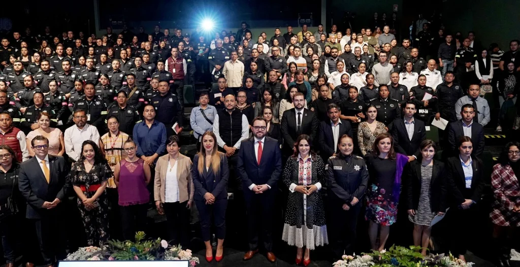 “Amplio auditorio con múltiples filas de personas reunidas en un evento institucional; en la primera fila hay asistentes de pie frente a una gran audiencia, y en filas posteriores se observan numerosas personas uniformadas, todo bajo iluminación interior con una luz brillante al fondo.”